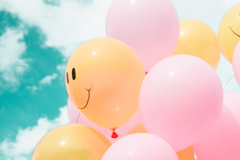 Photo of smiling colourful balloons against blue sky with fluffy clouds